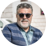 A headshot of a smiling man with a grey beard, wearing sunglasses and a blue striped cardigan.