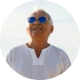 An older man with white hair and blue sunglasses, wearing a white shirt and looking upwards.
