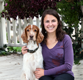 Person sitting with a dog in front of a white fence and greenery.
