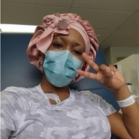 Person wearing a mask and pink headwrap, making a peace sign with hand.