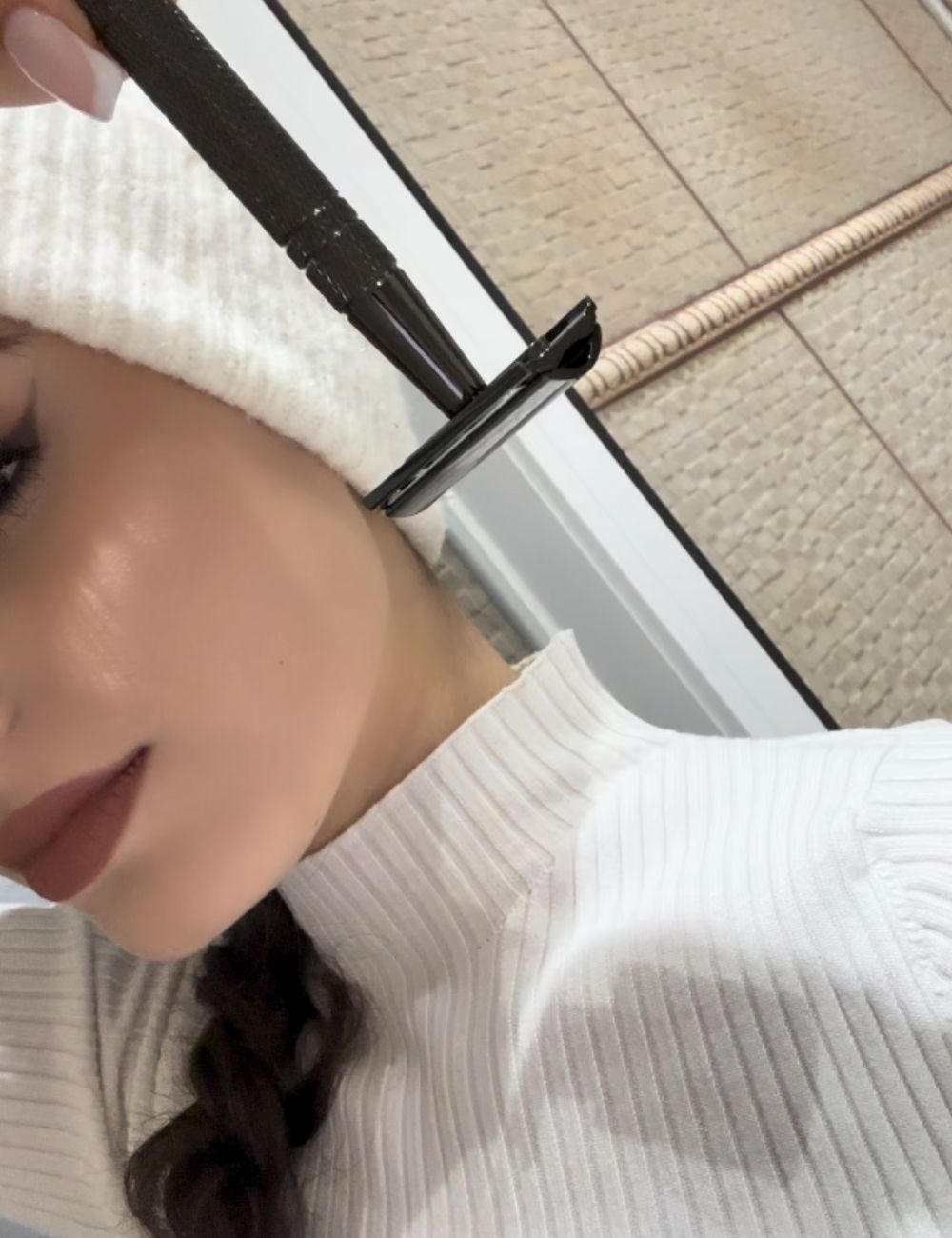 Person holding a razor near their face, wearing a white sweater and hat.