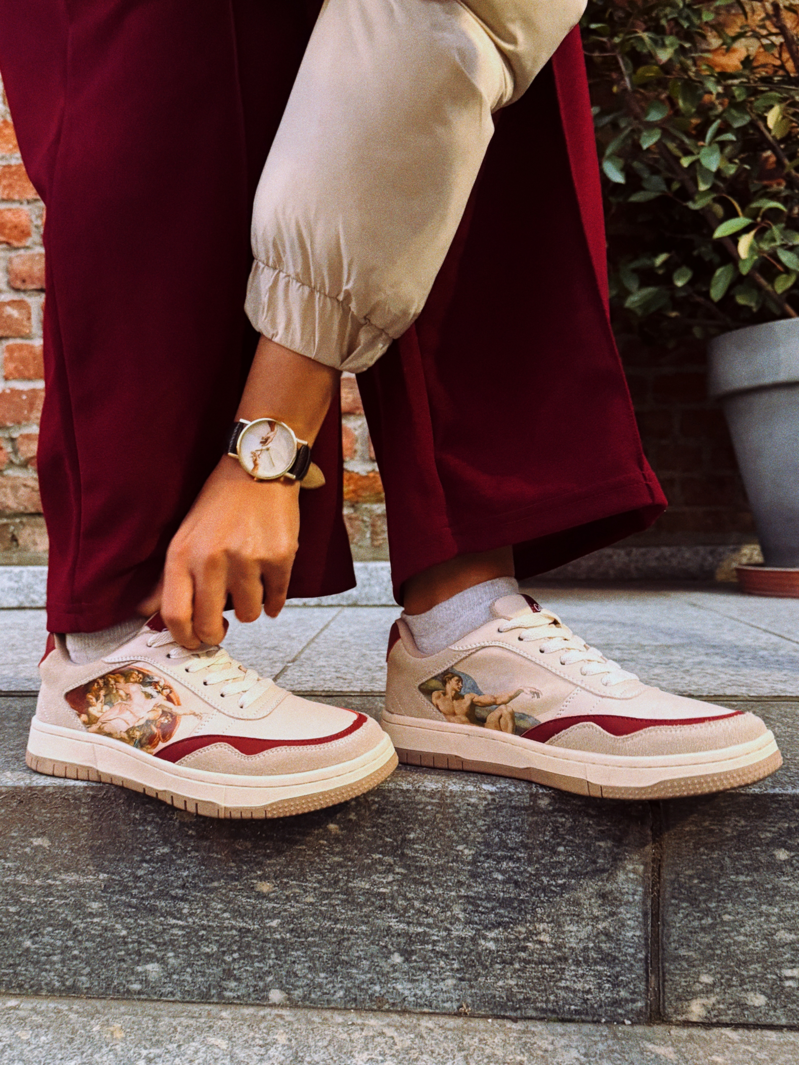 Person wearing sneakers with a Renaissance art design, burgundy pants, and a watch.