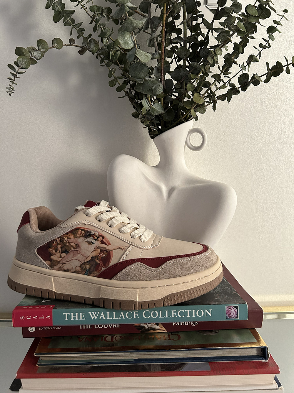 Sneaker with artistic design on books, next to a vase with green foliage.