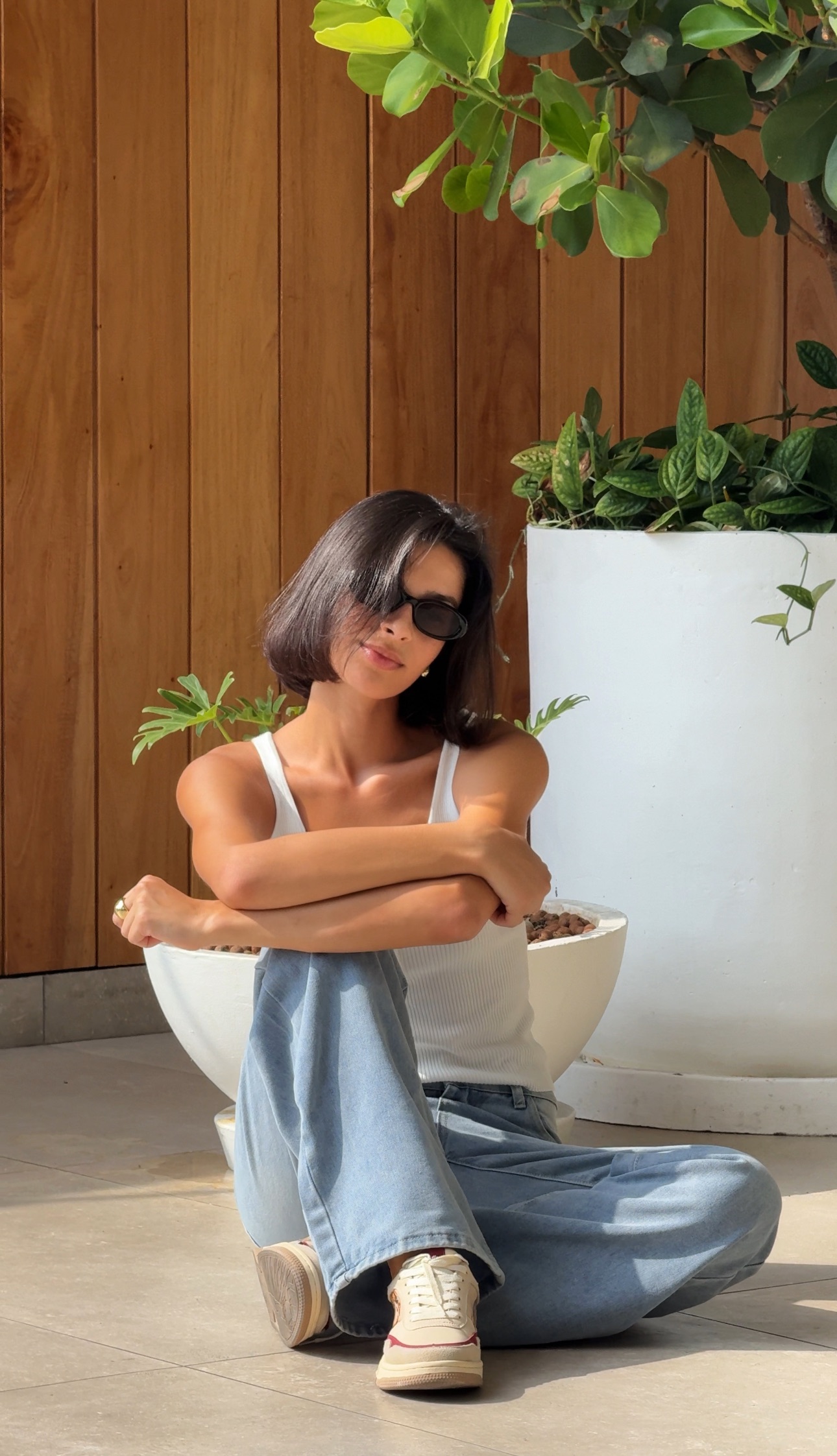 Person wearing sunglasses sitting near plants against a wooden wall.