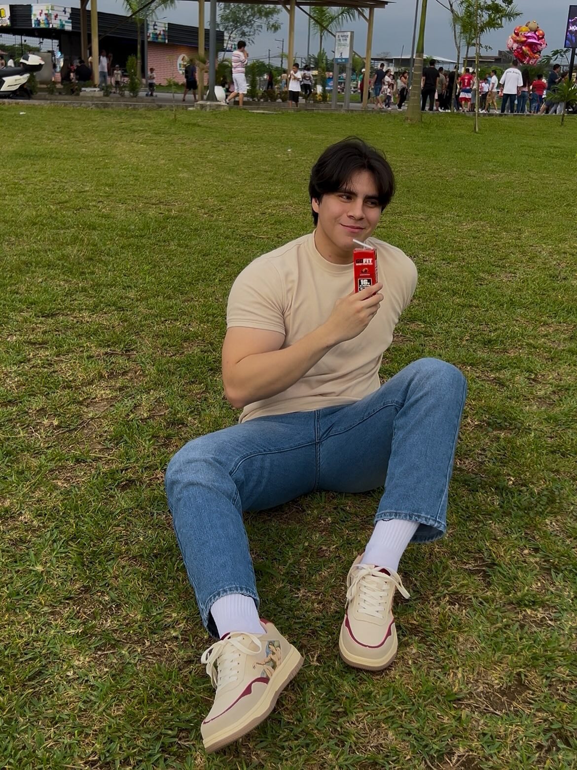 Person sitting on grass holding a red drink, with people in the background.