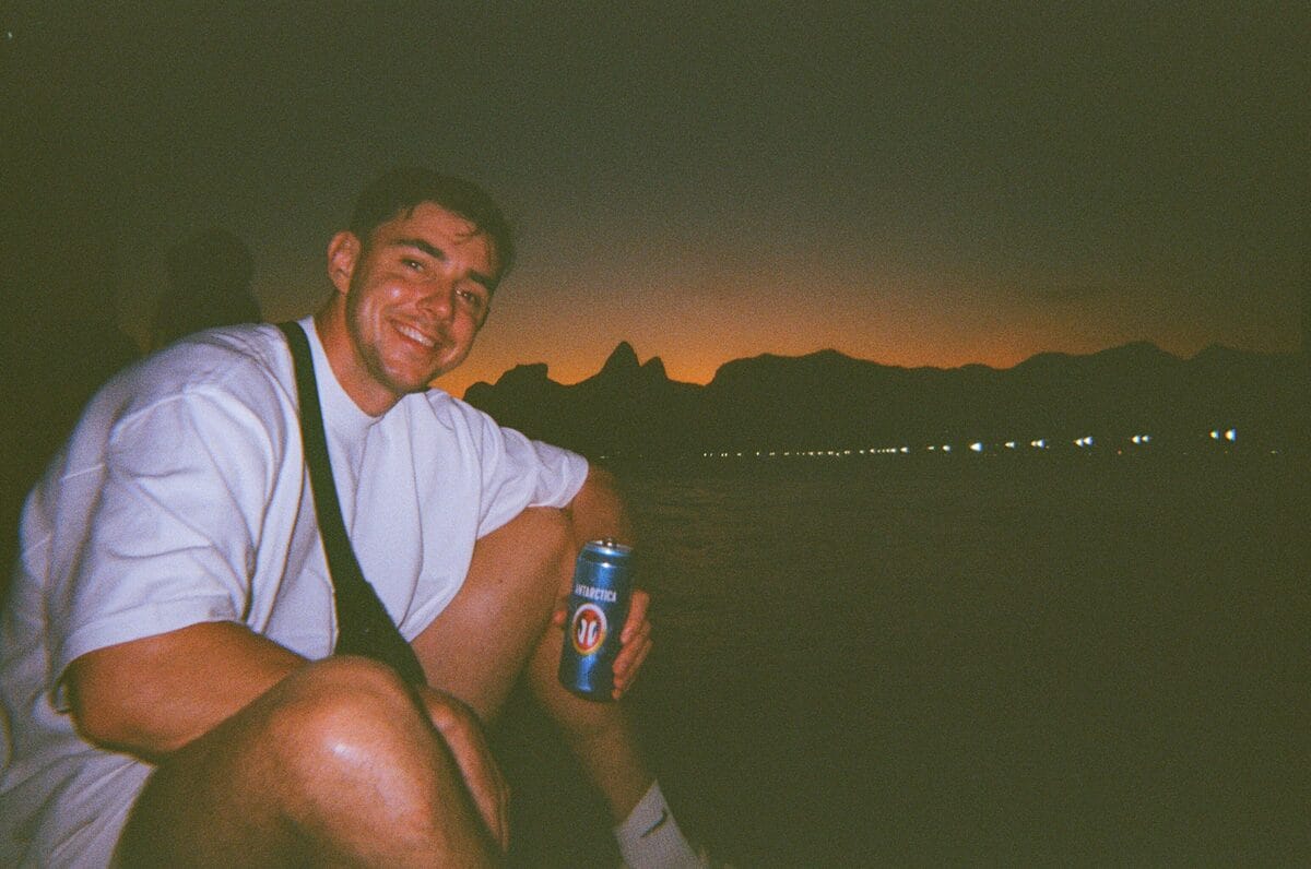 A man in a white shirt smiles at the camera, holding a can, with mountains silhouetted by a sunset.