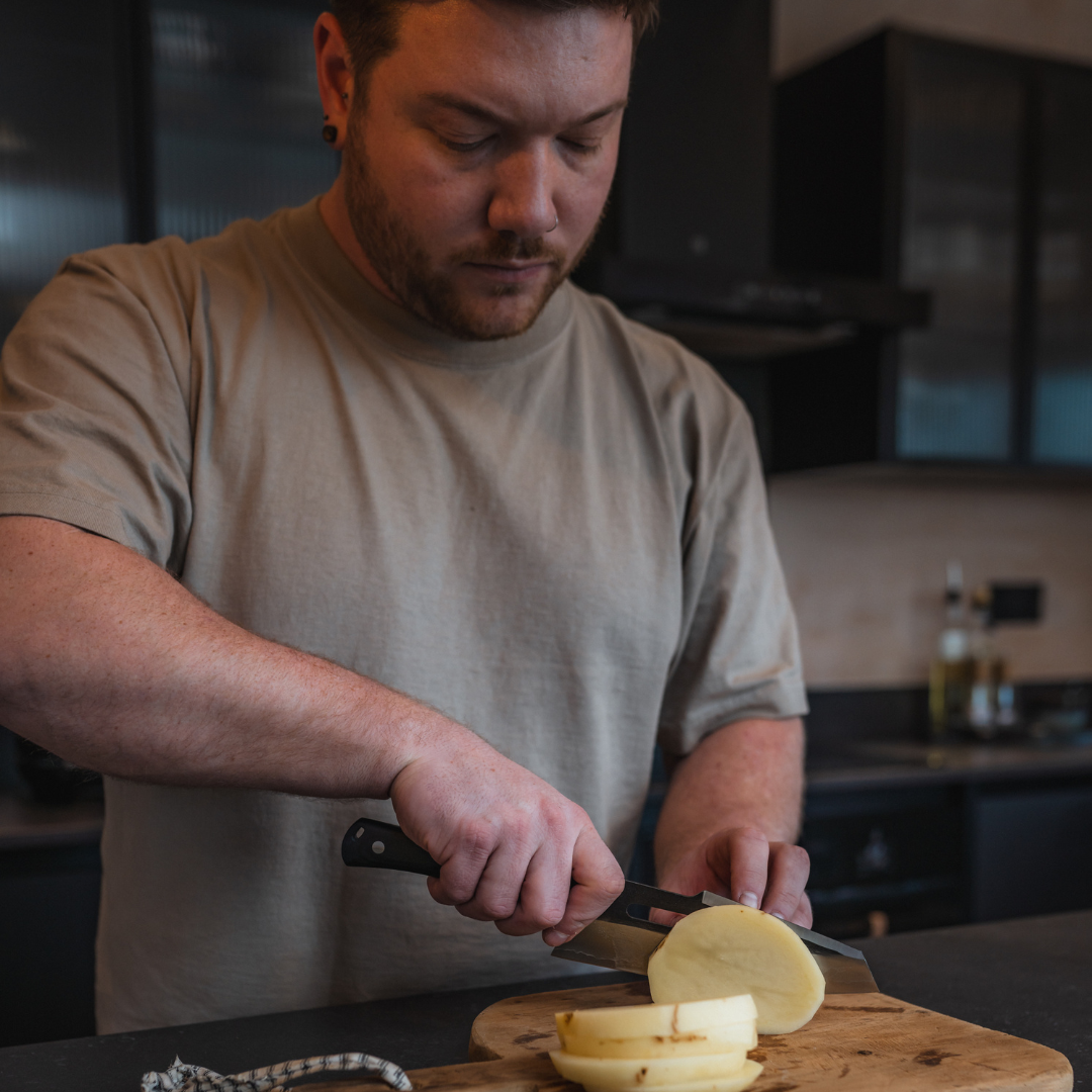 Person slicing a potato on a wooden cutting board in a kitchen.