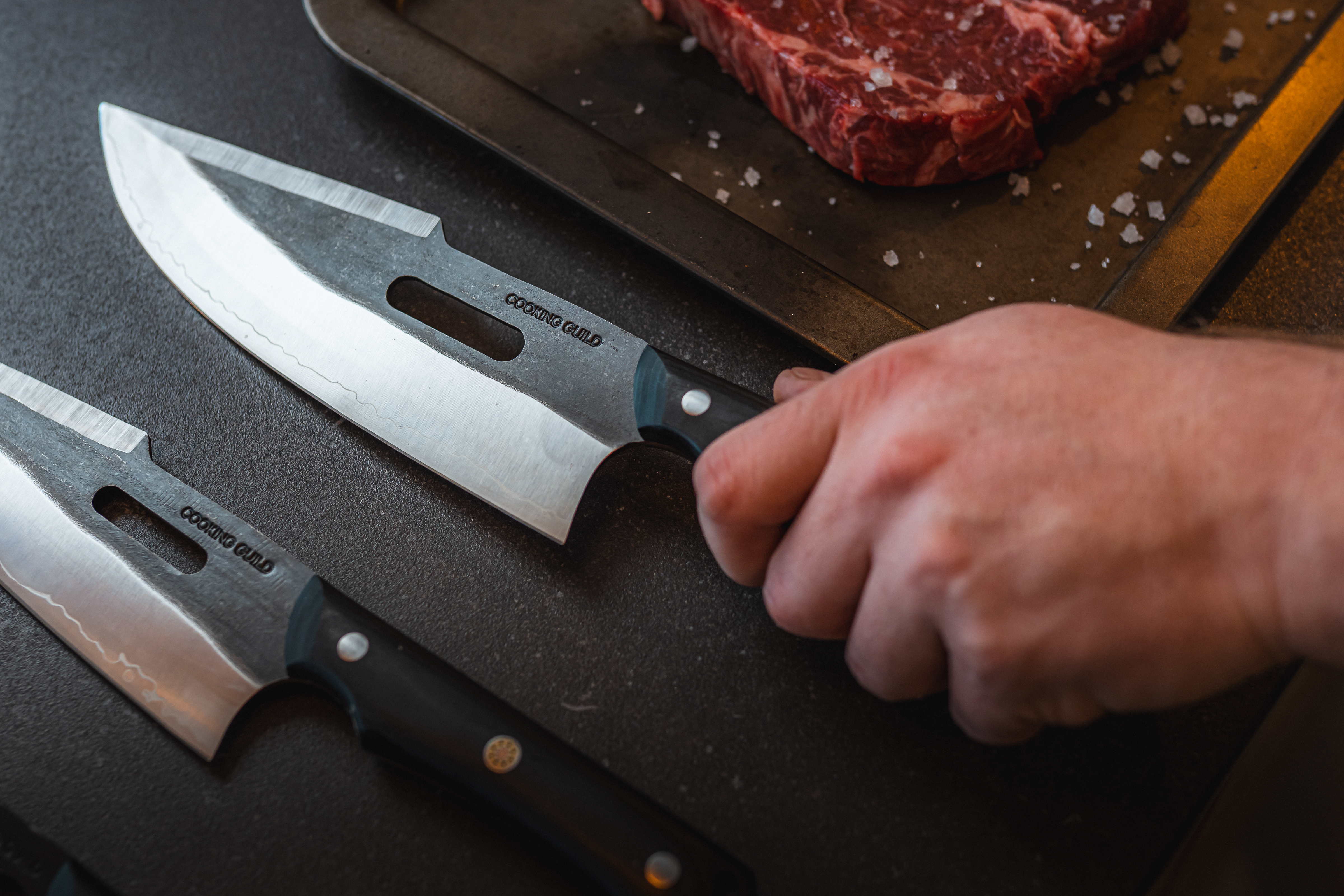 Hand holding a knife near a raw steak on a tray.