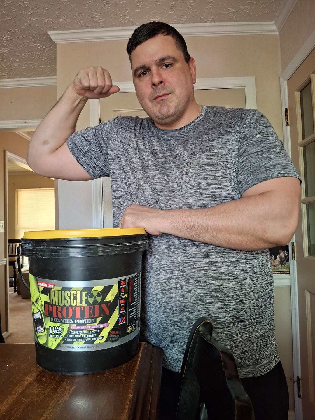 Man flexing arm next to a large container of protein powder.