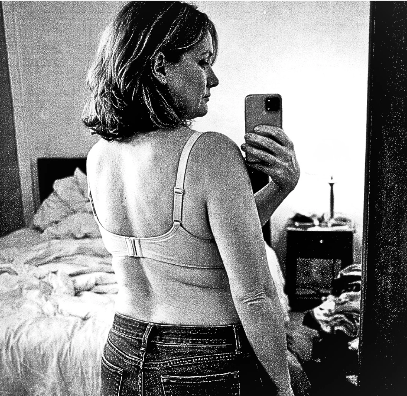 A black and white photo of a woman in a bra and jeans taking a selfie in a mirror.