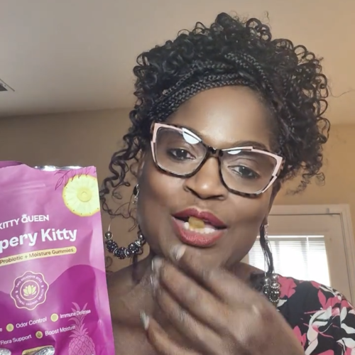 A woman with glasses and braided hair holds a purple bag of gummies while eating one.