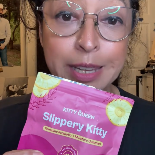 A woman wearing glasses holds up a pink pouch of Slippery Kitty feminine probiotic gummies.