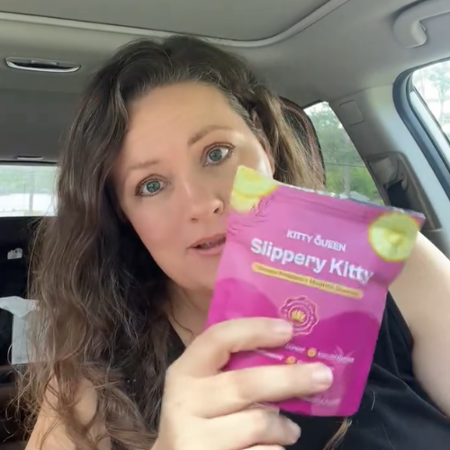 A woman with long brown hair sits in a car, holding up a pink pouch of Slippery Kitty.