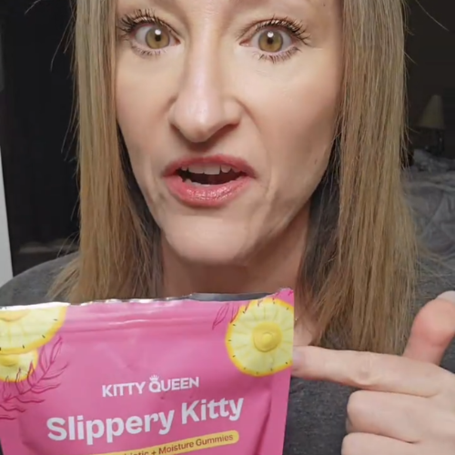 A woman with a surprised expression points to a pink pouch of Kitty Queen Slippery Kitty gummies.