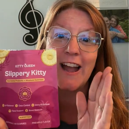 A smiling woman with glasses holds up a pink package of Slippery Kitty feminine probiotic gummies.