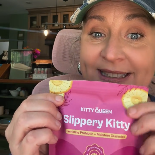 A woman wearing a cap smiles while holding a pink pouch of Kitty Queen Slippery Kitty gummies.
