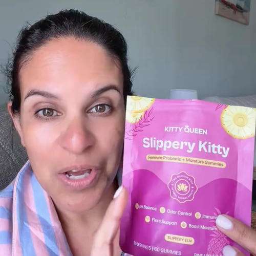 A woman holds up a pink pouch of Kitty Queen Slippery Kitty feminine probiotic gummies while talking.