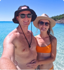 A couple in hats and sunglasses at a beach with clear blue water.