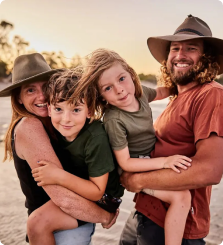Family of four smiling outdoors, wearing hats in a sunny setting.