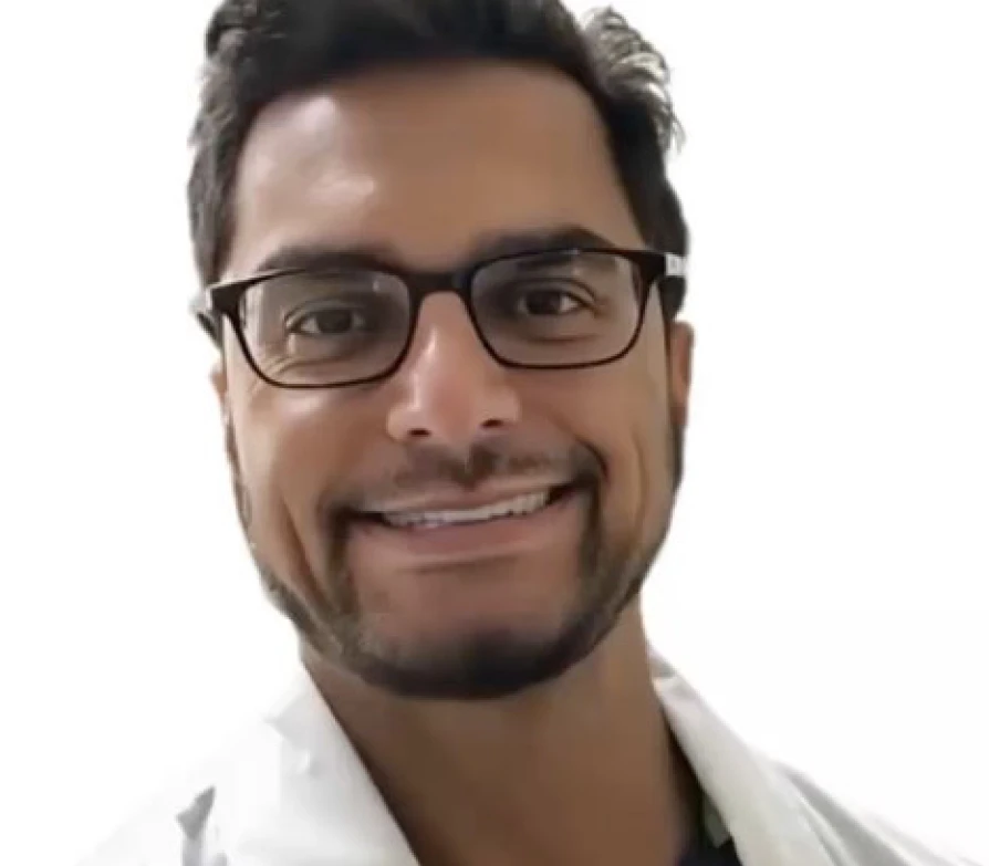 A headshot of a smiling man with dark hair, glasses, and a beard, wearing a white coat.