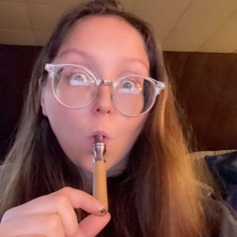 Person with glasses holding a wooden pipe to their mouth.