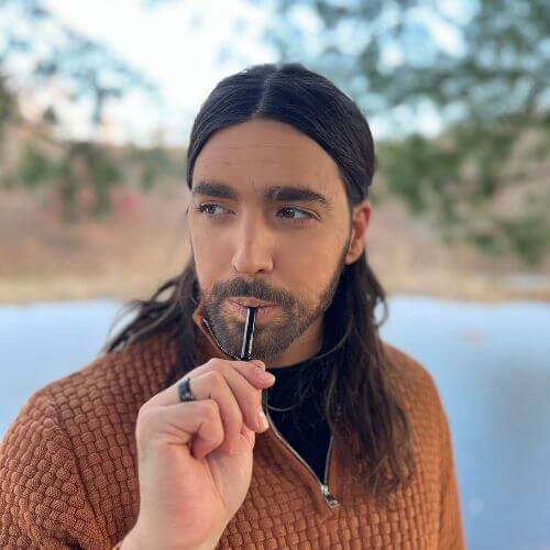 Person with long hair holding a pen near their mouth, outdoors.