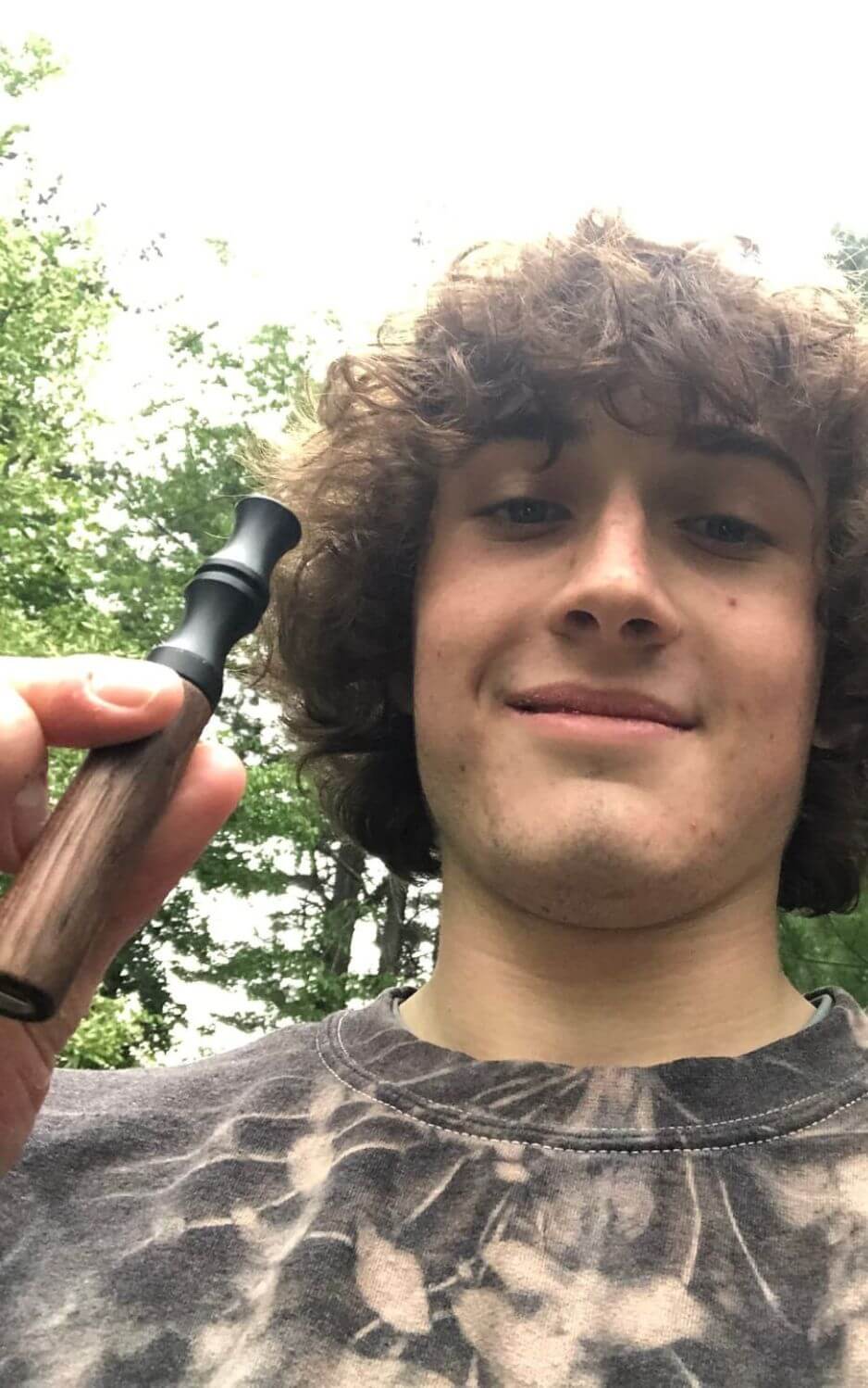 Person holding a wooden pipe outdoors, smiling at the camera.