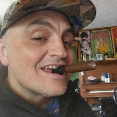 Person wearing a cap, holding a cigar in their mouth, with colorful artwork in the background.