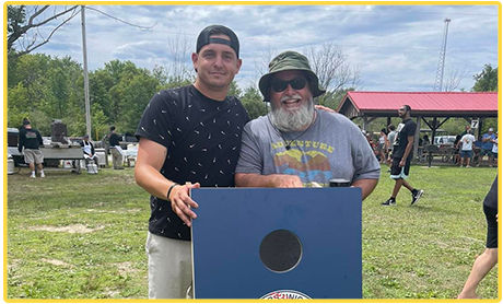 Two people smiling outdoors at a park, standing by a cornhole board.