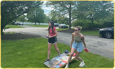 Two women playing cornhole in a grassy backyard setting.