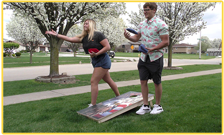 Two people playing cornhole in a park with blooming trees in the background.