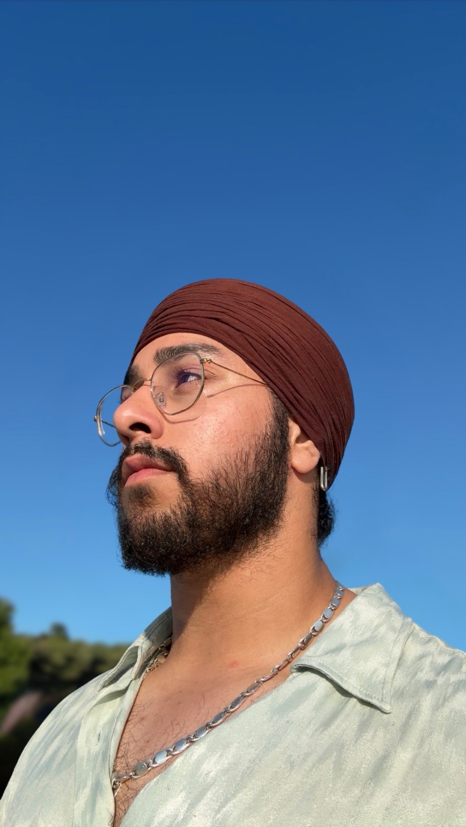 Person wearing glasses and a turban, looking up at the clear blue sky.