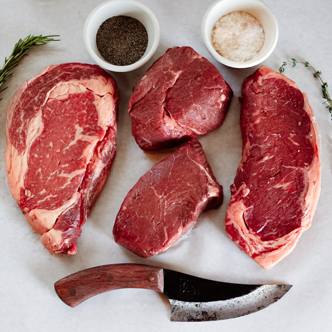 Raw steaks, herbs, salt, pepper, and a knife on a white surface.
