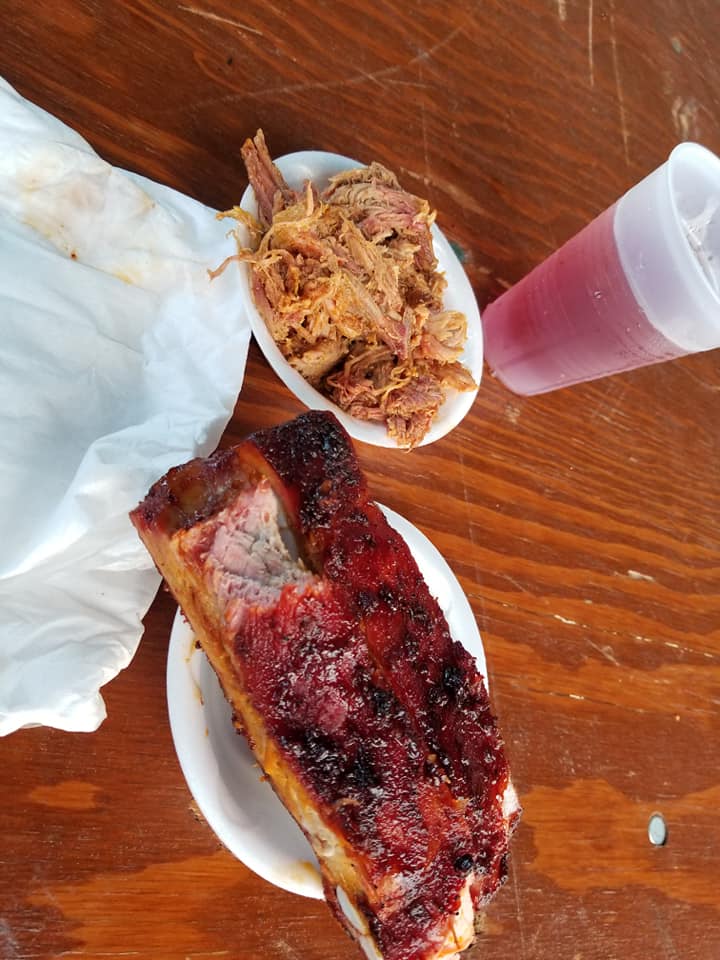Barbecue ribs, pulled pork in bowls, and a drink on a wooden table.