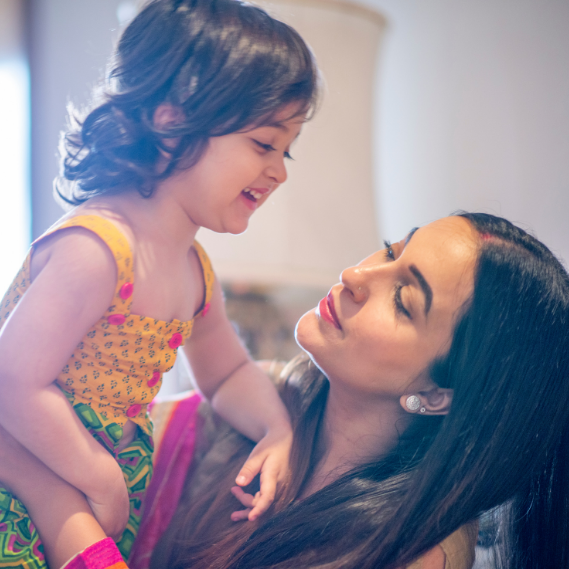 Woman holding a smiling child indoors, both looking at each other affectionately.
