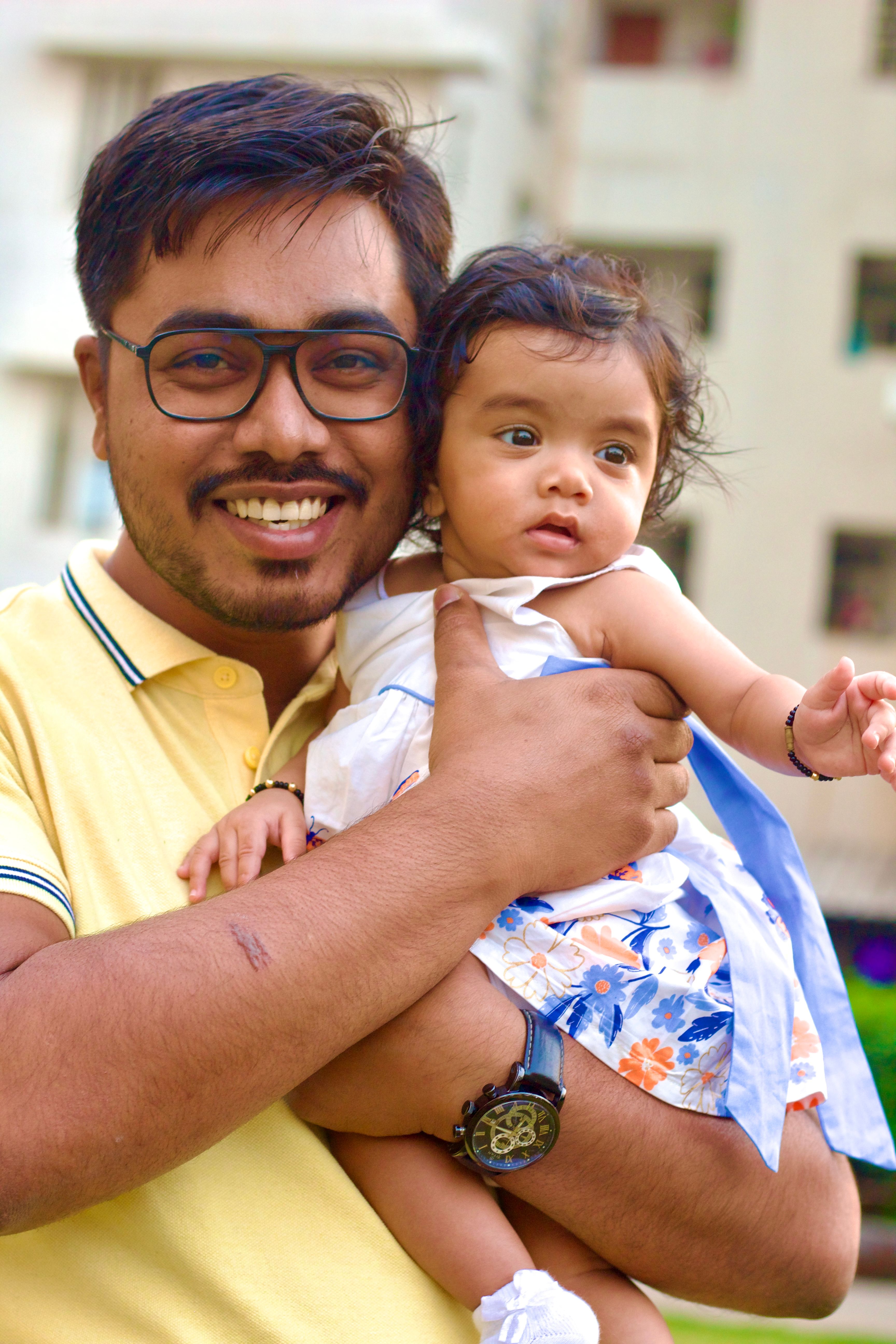 A smiling man with glasses in a yellow shirt holds a baby in a floral dress outdoors.
