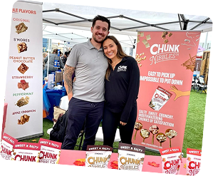 Two people standing at a booth displaying Chunk Nibbles snacks outdoors.