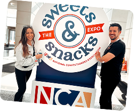 Two people posing beside a 'Sweets & Snacks Expo' sign indoors.