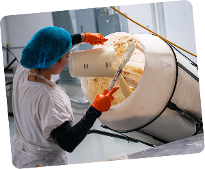 Person in a hairnet and gloves pouring liquid into a large container.