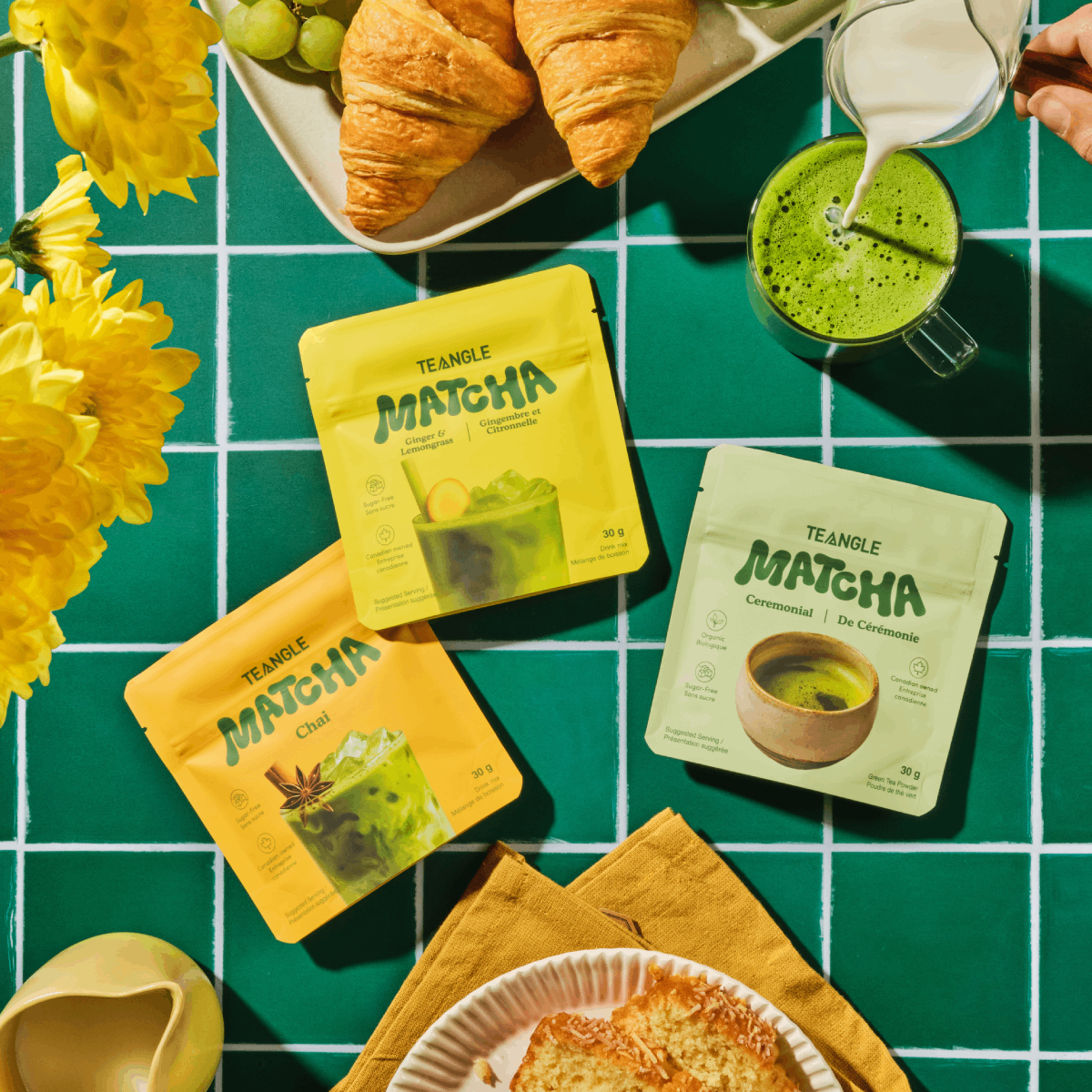 Assorted matcha packets and croissants on a green tiled surface with flowers.