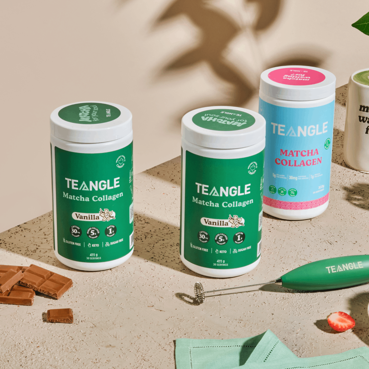 Three Teangle Matcha Collagen containers with mixer and chocolate pieces on a table.