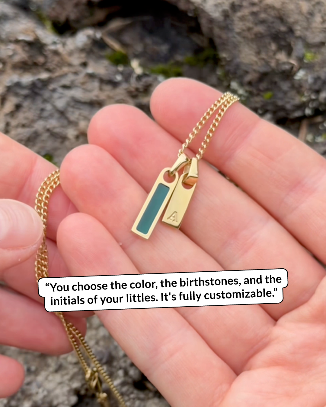 A hand holding a gold necklace with two small rectangular pendants, one teal and one with the initial A.