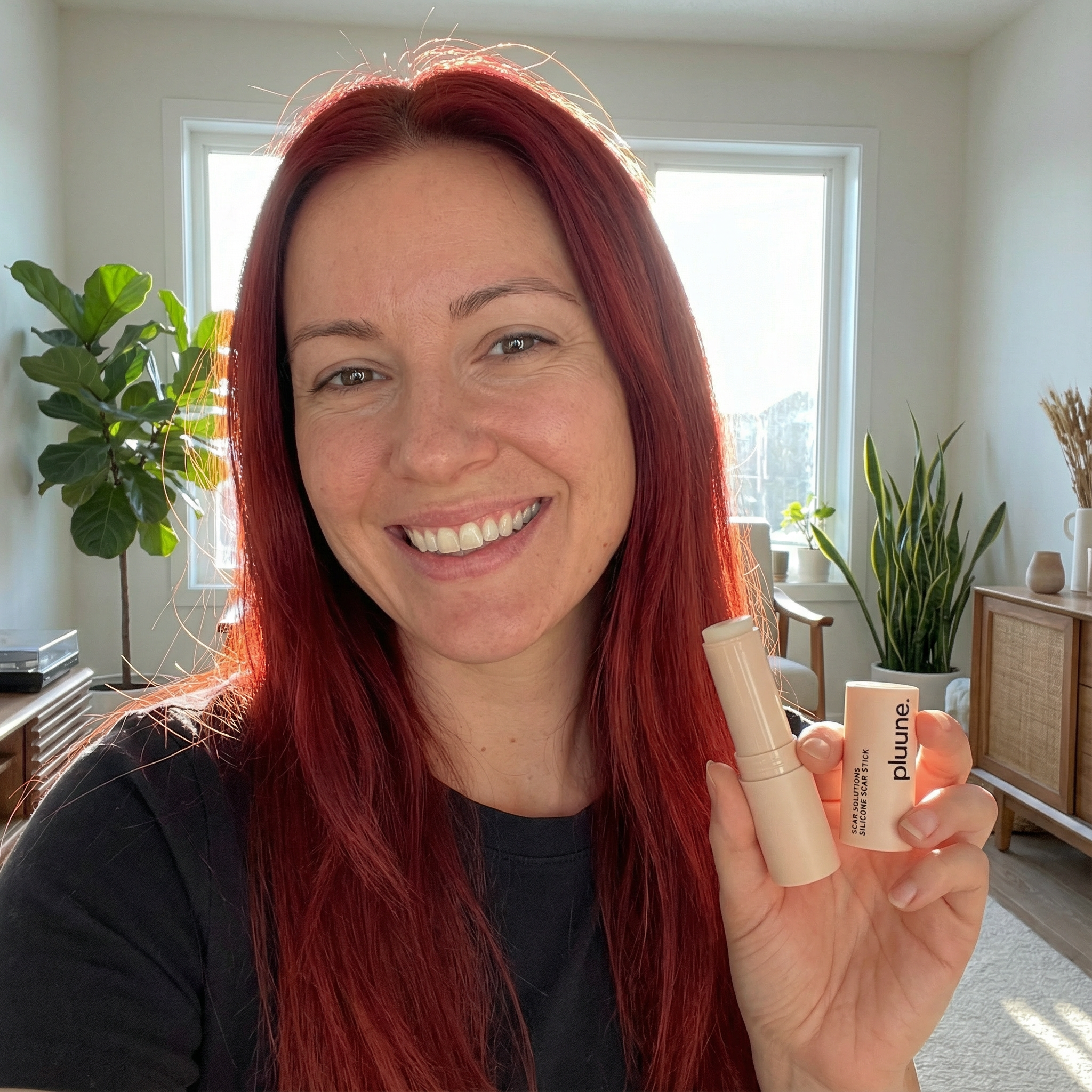 Person smiling and holding a makeup product in a well-lit room with plants.