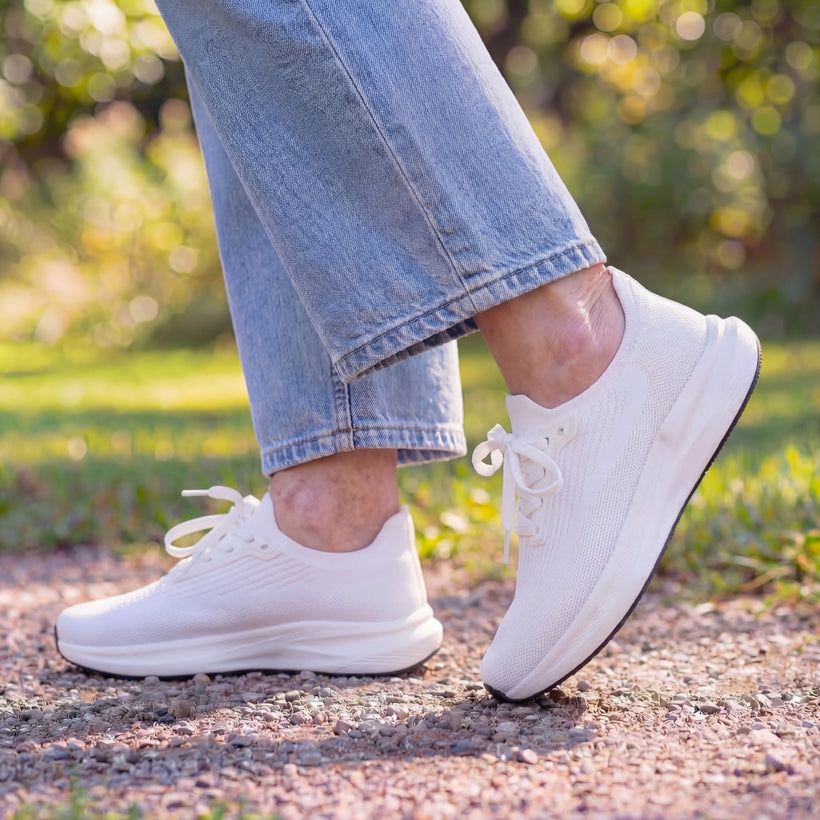 A close-up of a person wearing white sneakers and blue jeans walking on a gravel path.