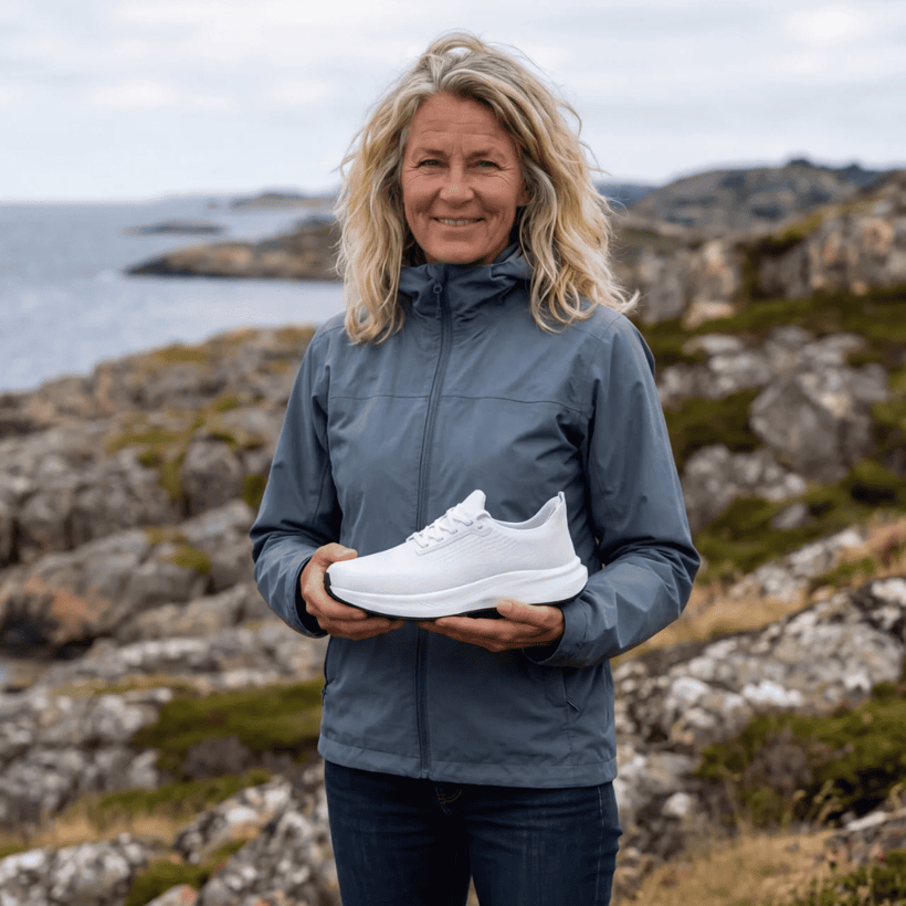 A woman in a blue jacket holds a white sneaker while standing on a rocky coast.