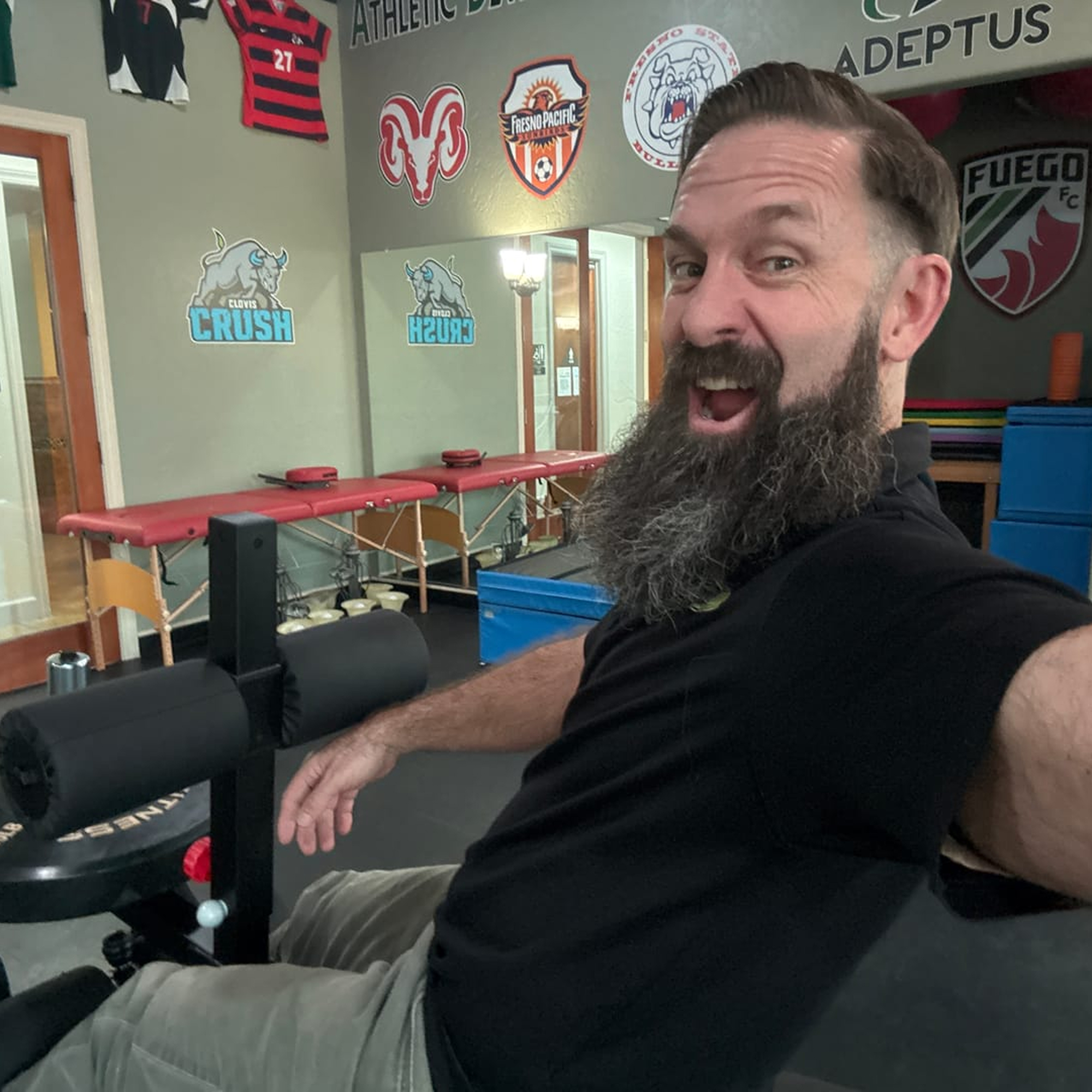 Man with a beard smiling in a gym with sports logos on the wall.
