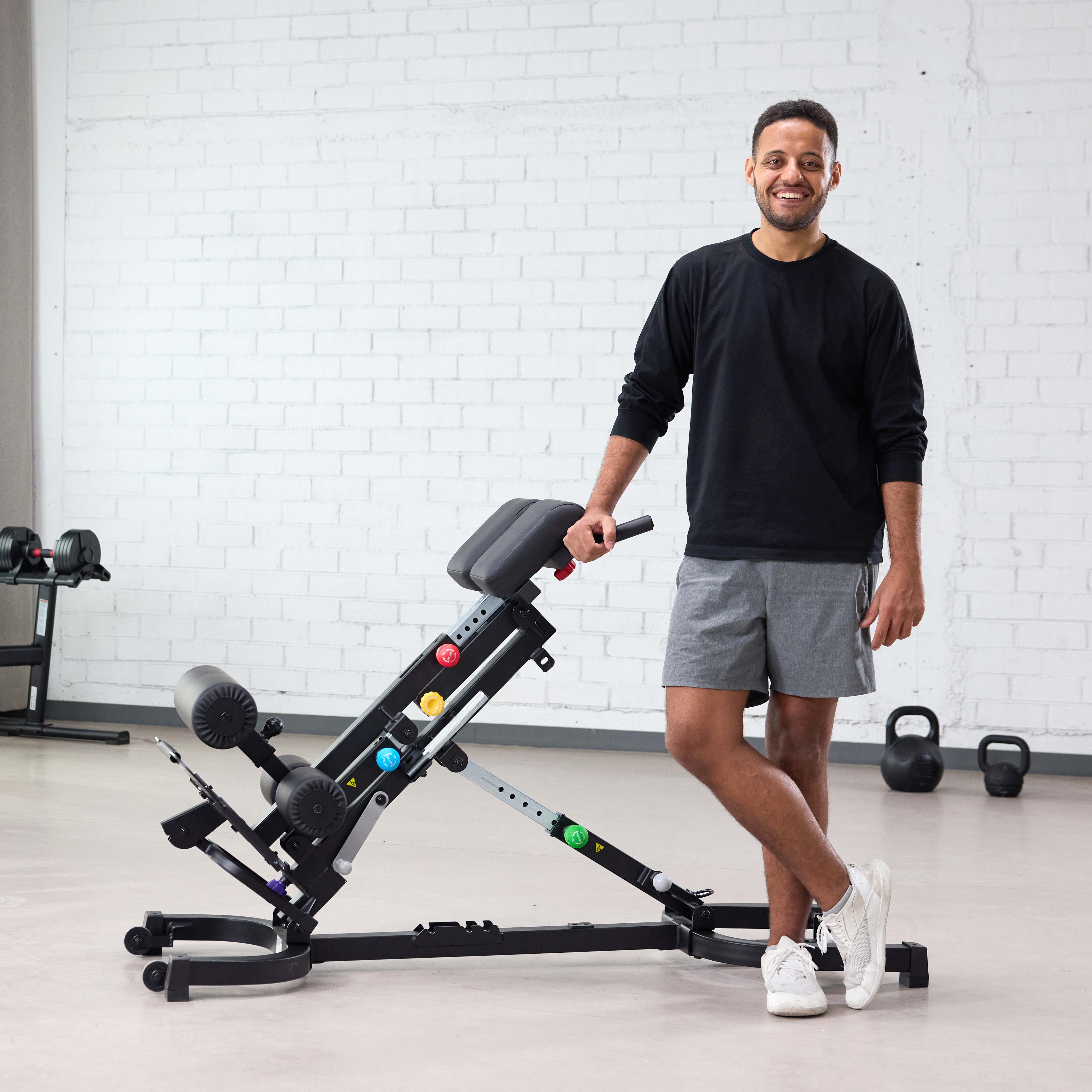 Man smiling, standing with an adjustable exercise bench in a gym setting.