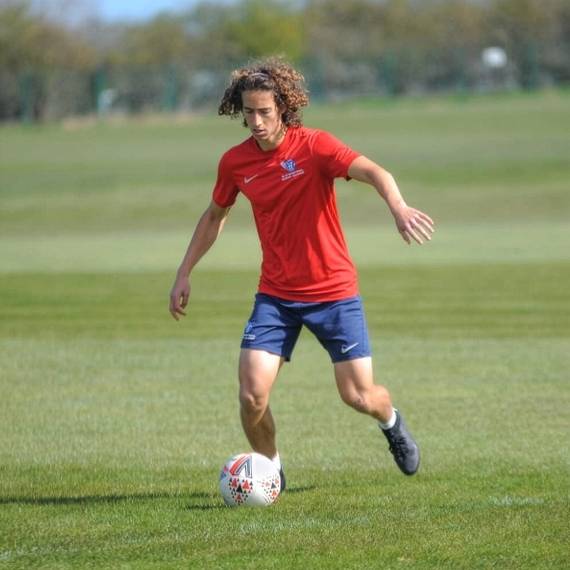 Person in a red shirt and blue shorts playing soccer on a grassy field.