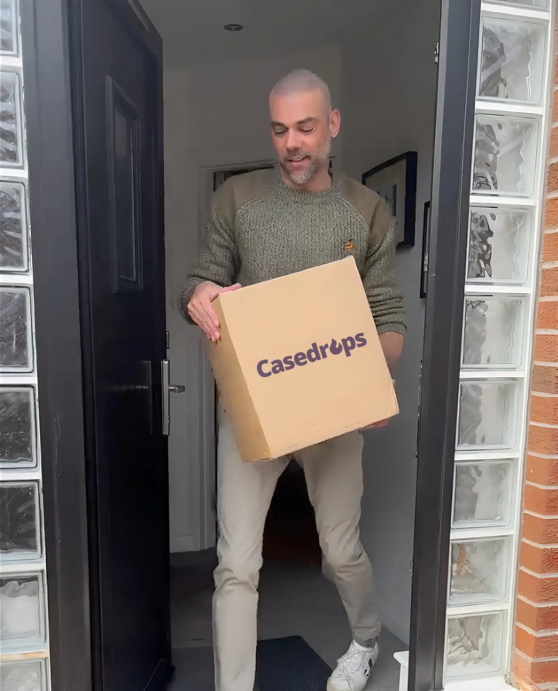 A man in a green sweater carries a cardboard box with the Casedrops logo through a doorway.