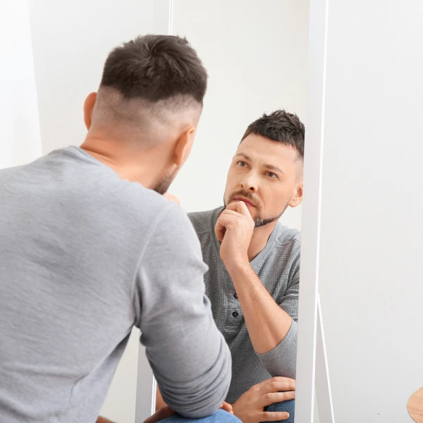 Man in gray shirt looking at himself in a mirror.